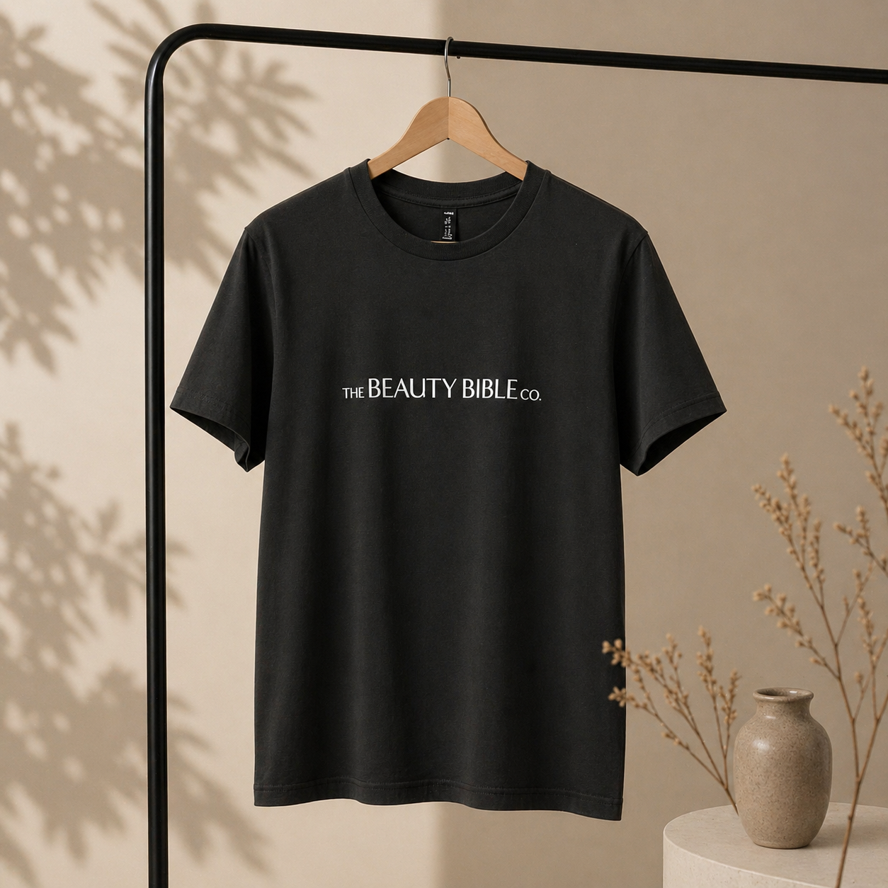 Black t-shirt with 'The Beauty Bible Co.' logo on a hanger against a beige wall.