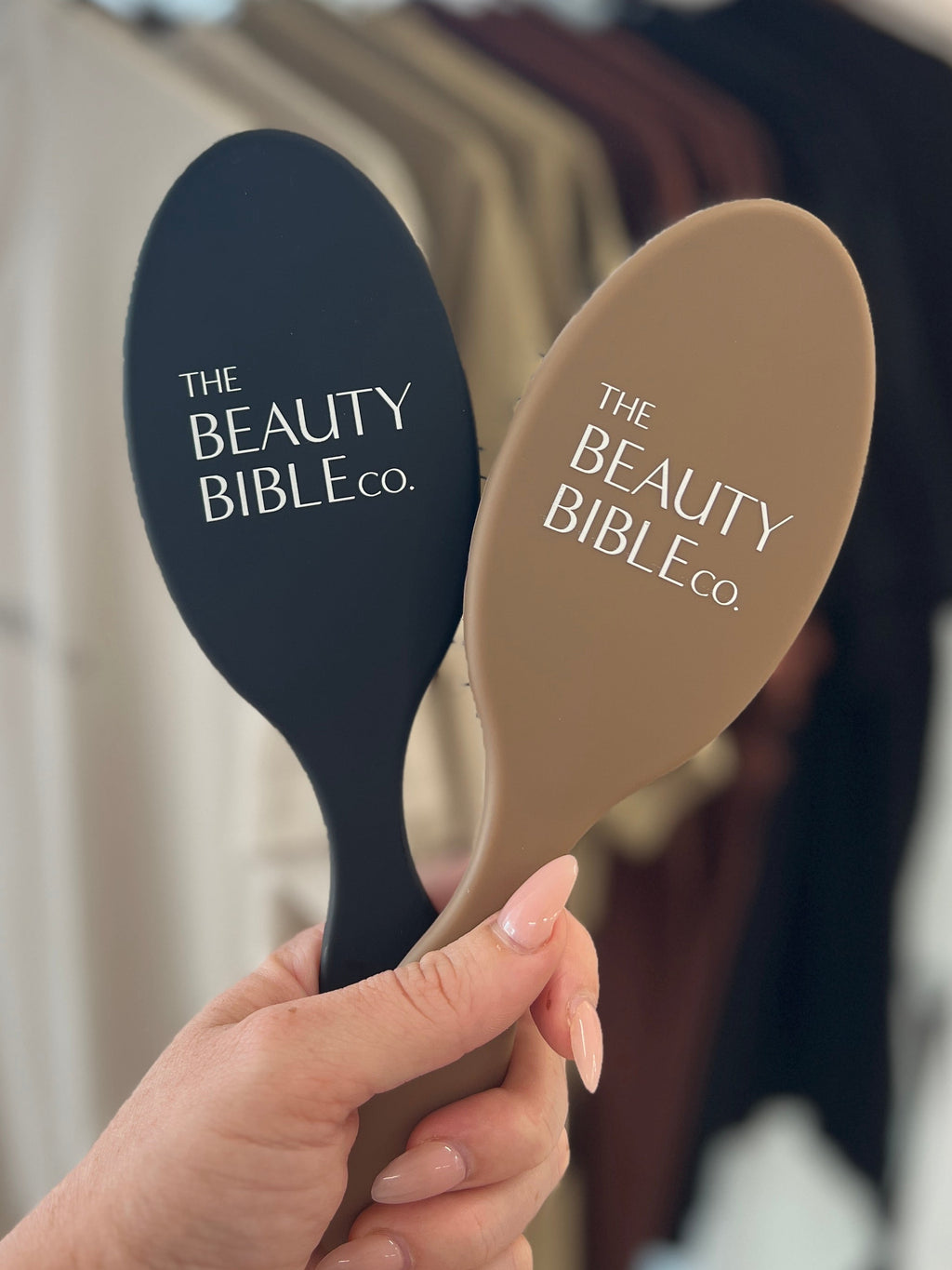 Two hairbrushes with 'The Beauty Bible Co.' branding held in front of a clothing rack.